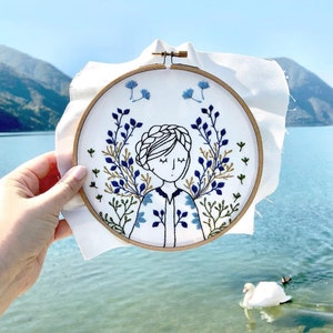May include: Embroidered hoop art featuring a woman with closed eyes surrounded by blue and green floral designs.