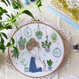 May include: Embroidered hoop art featuring a woman with brown hair holding a cat. The woman is surrounded by potted plants and flowers in a variety of colors. The hoop is white with a light blue background. The text "Tamar Nahir-Yanai" is visible in the lower left corner.