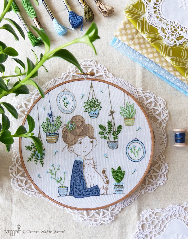 May include: Embroidered hoop art featuring a woman with brown hair holding a cat. The woman is surrounded by potted plants and flowers in a variety of colors. The hoop is white with a light blue background. The text "Tamar Nahir-Yanai" is visible in the lower left corner.