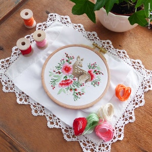 May include: A white embroidery hoop with a brown wooden frame. The hoop contains a hand-stitched design of a fawn standing in a field of red and pink flowers. The hoop is sitting on a white cloth with a lace border.