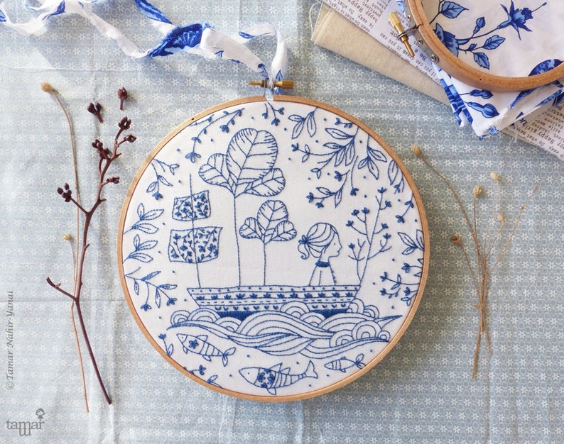 May include: Blue and white embroidery hoop featuring a design of a girl in a boat surrounded by trees and fish. The embroidery is on a white fabric background.