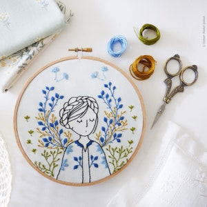 May include: Embroidered hoop art featuring a woman with closed eyes surrounded by blue and green floral designs. The hoop is made of wood and has a white fabric background.