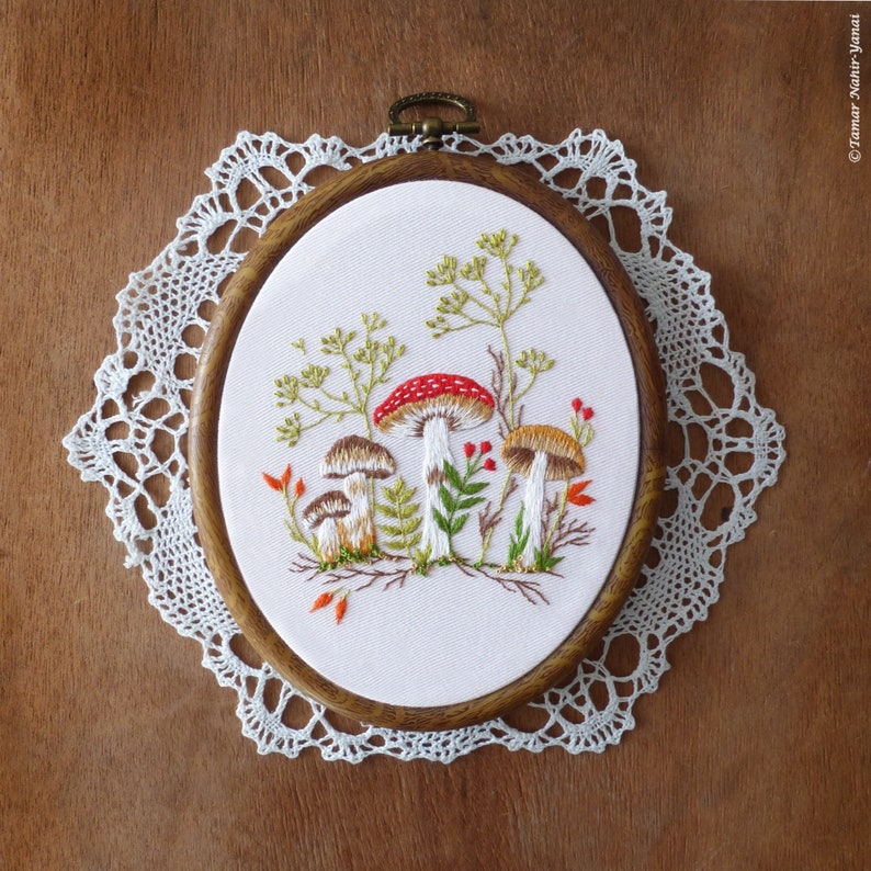 May include: Embroidered hoop art featuring a white background with three mushrooms, two brown and one red with white spots, surrounded by green leaves and red berries.
