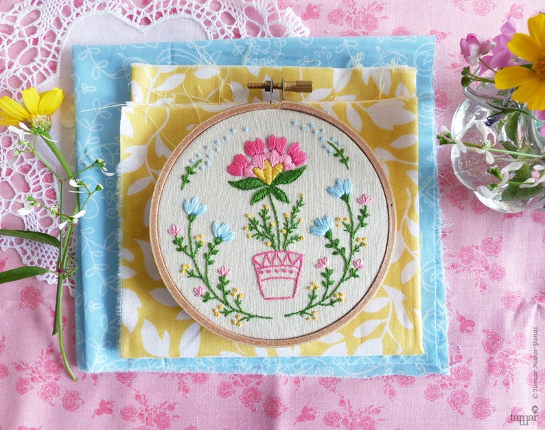 K&ouml;nnte beinhalten: Ein handgestickter Stickrahmen mit einem Blumenmuster. Der Rahmen liegt auf einem gelben Stoff mit einem wei&szlig;en Blumenmuster. Die Stickerei zeigt eine rosa Blume mit gr&uuml;nen Bl&auml;ttern in einem wei&szlig;en Topf.