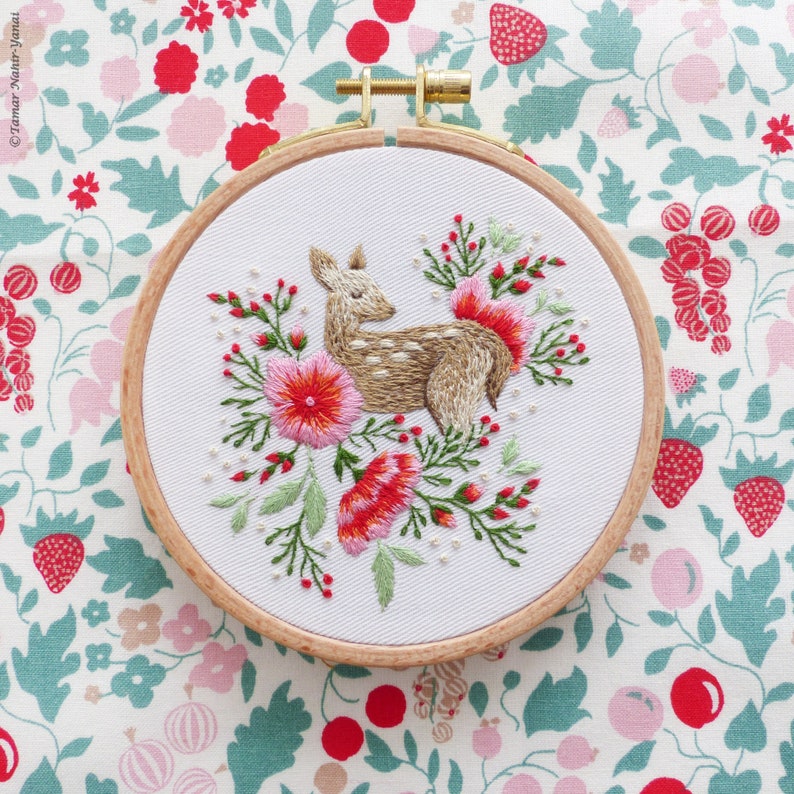 May include: Embroidered hoop art featuring a brown and white deer standing in a bed of pink and red flowers. The hoop is made of wood and has a white fabric background.