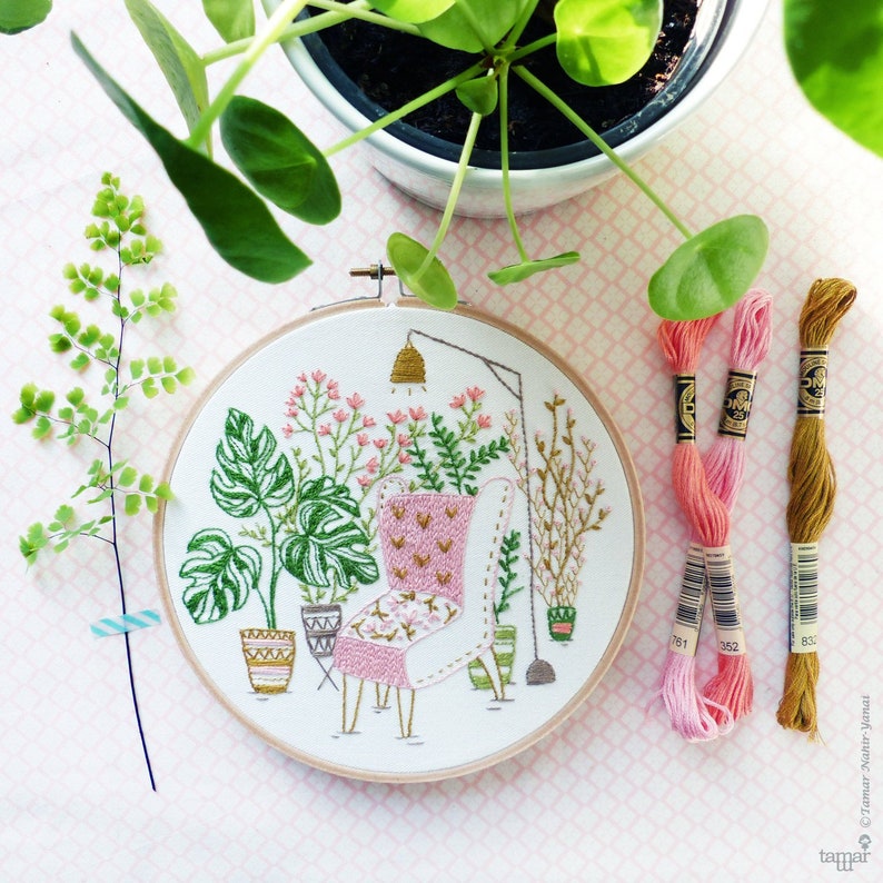 Puede incluir: Arte de bordado en aro que presenta un sill&oacute;n rosa con un patr&oacute;n floral, rodeado de plantas en macetas y una l&aacute;mpara colgante. El aro est&aacute; sobre un fondo estampado rosa y blanco con carretes de hilo de bordar.