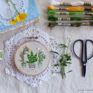 May include: An embroidery hoop featuring a botanical design of potted plants, surrounded by embroidery floss in various shades of green, yellow, and white. A pair of black scissors, a spool of green thread, and lace doilies complete the craft-themed arrangement.