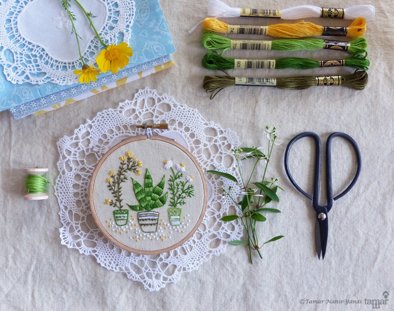 May include: An embroidery hoop featuring a botanical design of potted plants, surrounded by embroidery floss in various shades of green, yellow, and white. A pair of black scissors, a spool of green thread, and lace doilies complete the craft-themed arrangement.