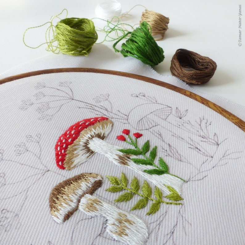 May include: Embroidered hoop art featuring two mushrooms with red and white caps, green leaves, and red berries on a white fabric background.