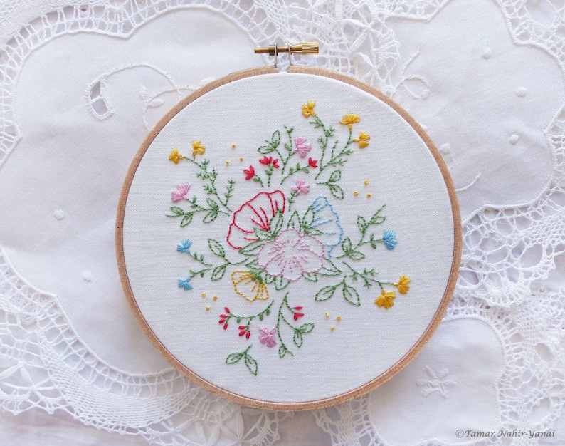 以下が含まれることがあります： 白い刺繍枠にカラフルな花の模様が施されています。花はピンク、赤、黄色、青です。模様は緑色の糸で刺繍されています。