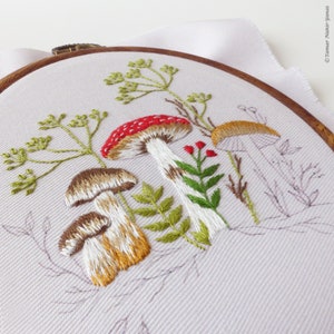 May include: Embroidered hoop art featuring a whimsical forest scene with mushrooms, ferns, and delicate greenery. The mushrooms are depicted in shades of red, brown, and white, with intricate details in the stitching.
