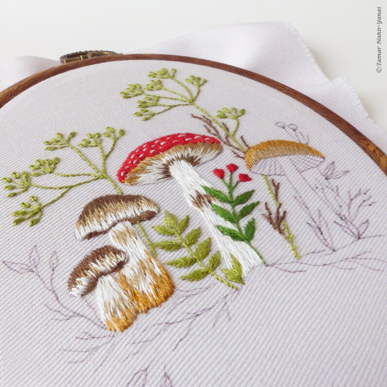 May include: Embroidered hoop art featuring a whimsical forest scene with mushrooms, ferns, and delicate greenery. The mushrooms are depicted in shades of red, brown, and white, with intricate details in the stitching.