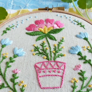 K&ouml;nnte beinhalten: Stickerei auf einem Stickrahmen mit einer rosa Blume in einem rosa Topf mit gr&uuml;nen Bl&auml;ttern und gelben Bl&uuml;ten. Der Hintergrund ist ein hellbeiger Stoff.
