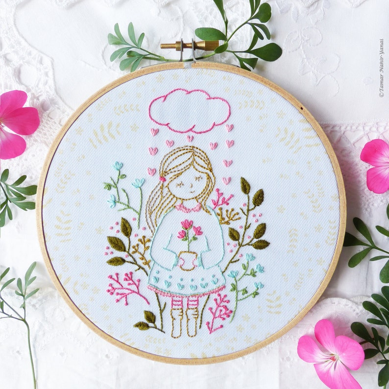May include: Embroidered hoop art featuring a girl with blonde hair holding a pot of pink flowers. The girl is standing in a field of green and blue flowers with a pink cloud above her head. Pink hearts are falling from the cloud.