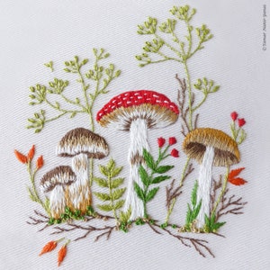 May include: Embroidered design of mushrooms and greenery on a white fabric background. The mushrooms are in various sizes and colors, including red, brown, and white. The greenery is in shades of green.