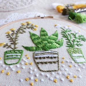 May include: Embroidered floral design featuring three potted plants in a wooden hoop. The plants include yellow flowers, a succulent, and daisies, all in green and white pots. The background is a light beige fabric with scattered yellow and white thread accents.