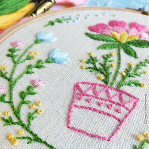 K&ouml;nnte beinhalten: Stickerei in einem Stickrahmen mit einer rosa Blume mit gelbem Zentrum in einem rosa Topf mit geometrischem Muster. Die Blume ist umgeben von gr&uuml;nen Bl&auml;ttern und kleinen rosa und gelben Blumen.
