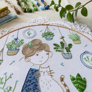 May include: Embroidered hoop art featuring a woman with brown hair and a cat, surrounded by hanging plants in blue and green pots. The background is white fabric with blue and green floral embroidery.