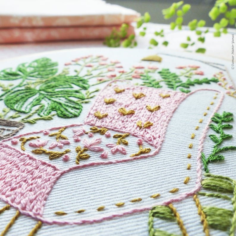 Puede incluir: Primer plano de una regadera bordada en rosa y dorado con hojas y flores verdes. El bordado se realiza sobre una tela blanca.