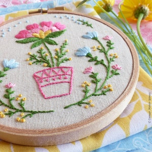 K&ouml;nnte beinhalten: Stickerei auf einem Stickrahmen mit einer rosa und gelben Blume in einem rosa Topf mit gr&uuml;nen Bl&auml;ttern und Stielen. Der Rahmen ist von einem Kranz aus kleinen gelben und blauen Blumen umgeben.
