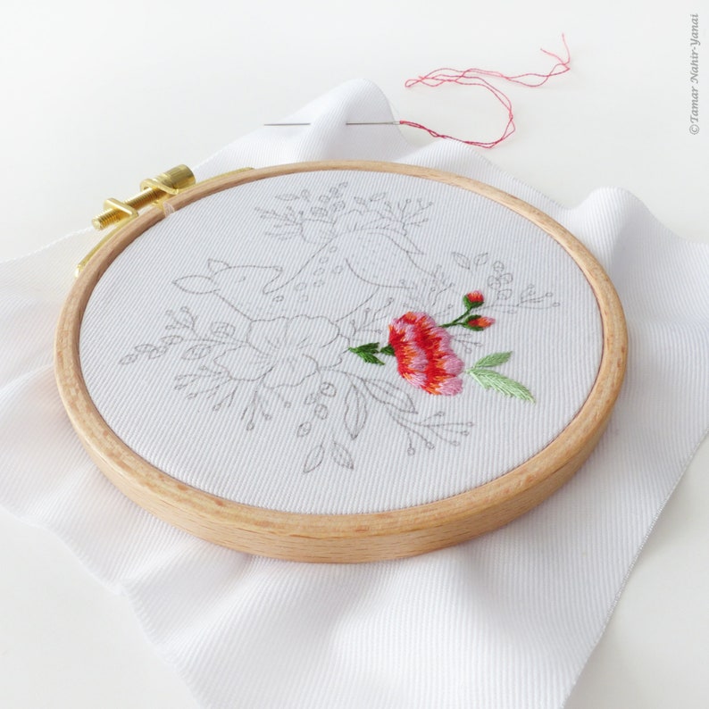 May include: A close-up of a white embroidery hoop with a floral design. The design includes a pink flower with green leaves and a few smaller pink flowers. The hoop is on a white fabric background.