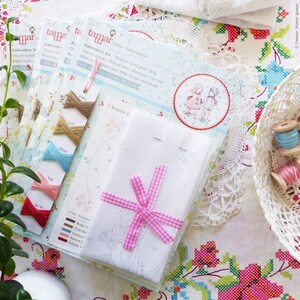May include: Embroidery kit with a printed fabric, illustrated color and stitch guide, DMC threads and needle. The kit includes a white fabric with a pink gingham ribbon and a printed design of a winter girl and boy. The text on the packaging reads "Tamar Nahir-Yanai Embroidery Kit Winter Girl and Boy".
