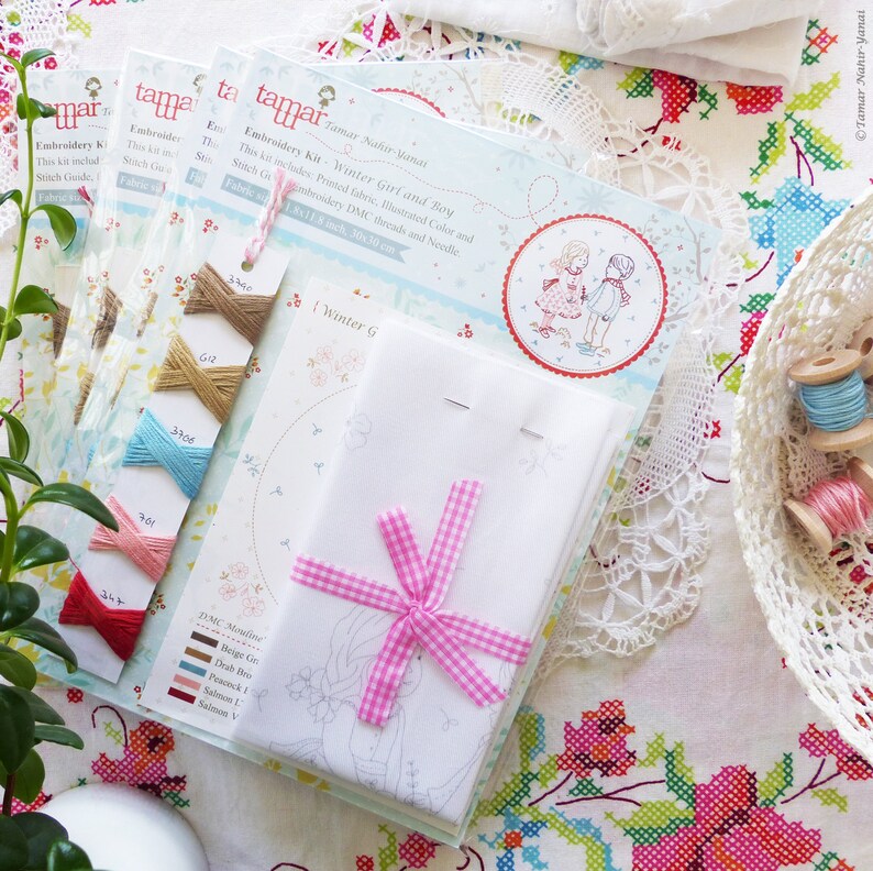 May include: Embroidery kit with a printed fabric, illustrated color and stitch guide, DMC threads and needle. The kit includes a white fabric with a pink gingham ribbon and a printed design of a winter girl and boy. The text on the packaging reads "Tamar Nahir-Yanai Embroidery Kit Winter Girl and Boy".
