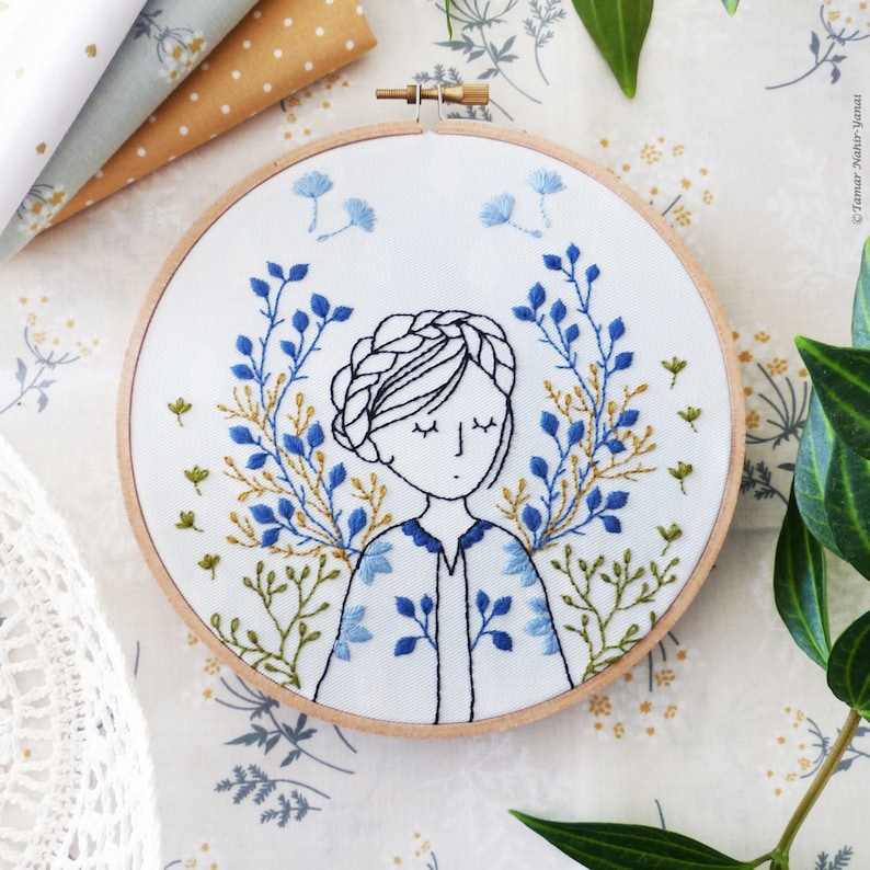 May include: Embroidered hoop art featuring a woman with closed eyes surrounded by blue and green floral designs.