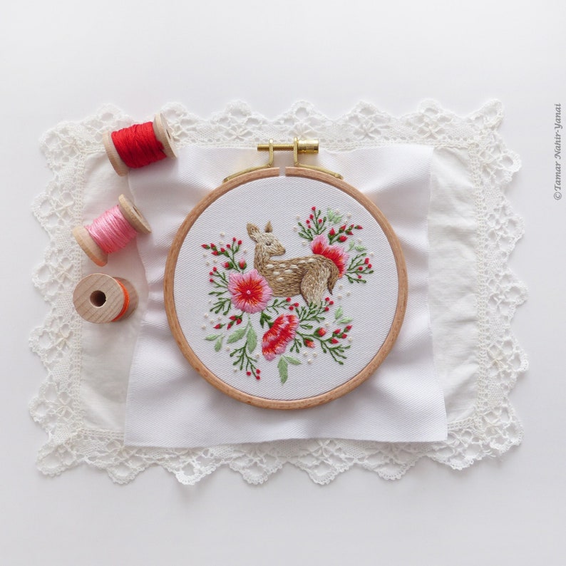 May include: A white embroidery hoop with a brown wooden frame. The hoop features a hand-stitched design of a fawn surrounded by pink and red flowers. The hoop is sitting on a white fabric with a lace trim.