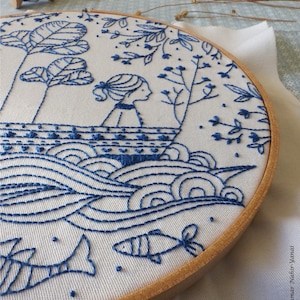 May include: Blue and white embroidery hoop featuring a design of a girl in a boat on a wavy sea with fish swimming below. The design is stitched onto a white fabric background.