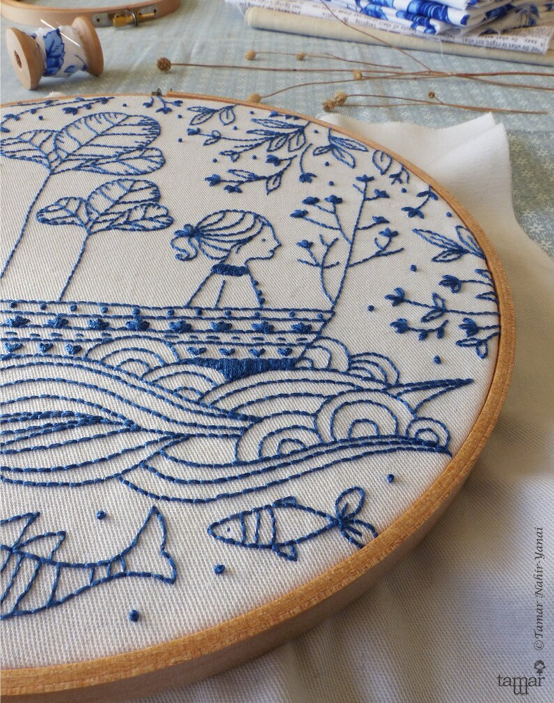 May include: Blue and white embroidery hoop featuring a design of a girl in a boat on a wavy sea with fish swimming below. The design is stitched onto a white fabric background.