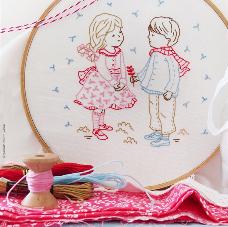 May include: Embroidered hoop art featuring two children, a girl in a pink dress and a boy in a blue shirt and red scarf, standing in a field of blue flowers. The hoop is made of wood and has a white fabric background.