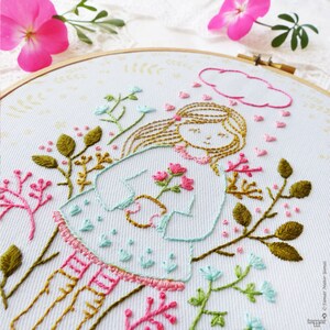 May include: Embroidered hoop art featuring a girl with brown hair holding pink flowers, surrounded by green leaves and pink flowers. The girl is wearing a light blue dress with a pink collar and pink and white striped stockings. A pink cloud with pink hearts is above her head.