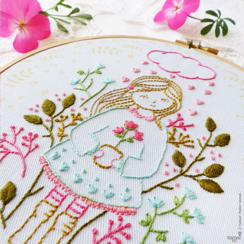 May include: Embroidered hoop art featuring a girl with brown hair holding pink flowers, surrounded by green leaves and pink flowers. The girl is wearing a light blue dress with a pink collar and pink and white striped stockings. A pink cloud with pink hearts is above her head.