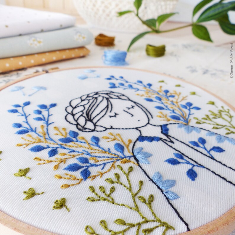 May include: Embroidered hoop art featuring a stylized illustration of a person with closed eyes surrounded by blue, green, and gold floral embroidery.