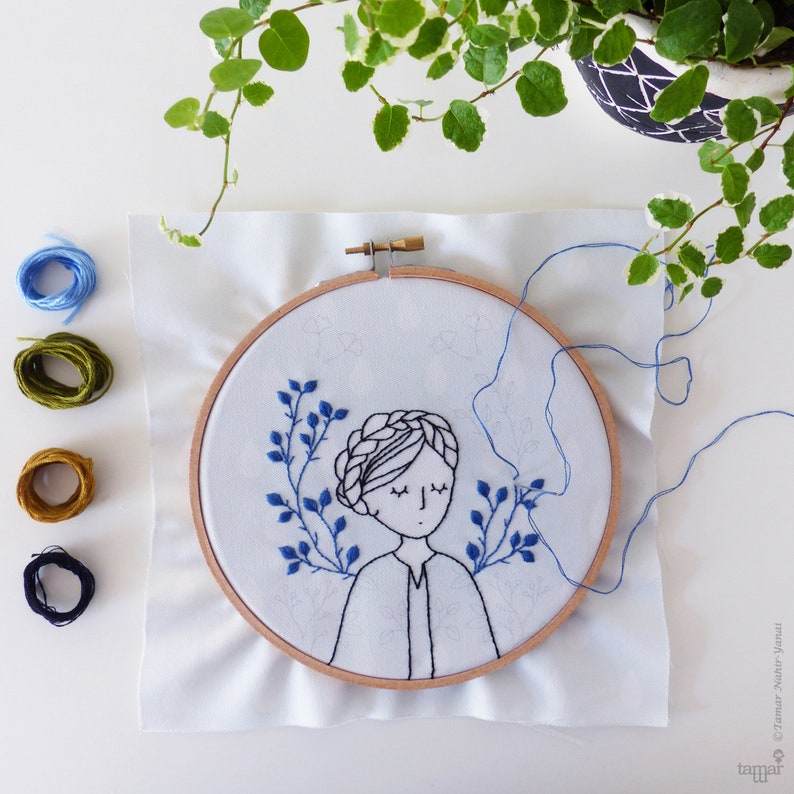 May include: A close-up of a hoop with embroidery of a woman with her eyes closed and a braid in her hair. The woman is surrounded by blue embroidered leaves. The hoop is on a white fabric background.