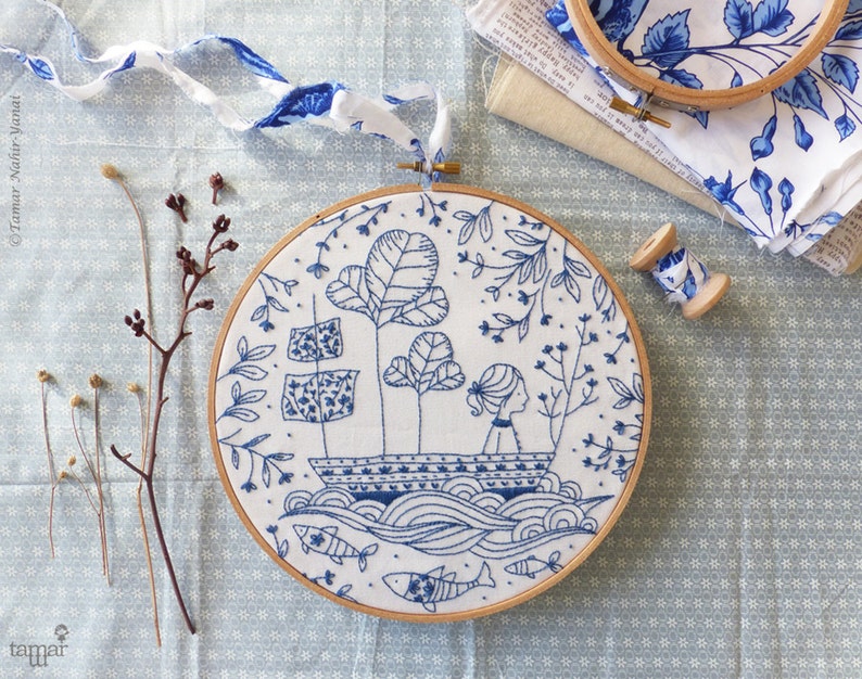 May include: Blue and white embroidery hoop featuring a design of a girl in a boat surrounded by flowers and fish. The hoop is on a blue and white patterned fabric.