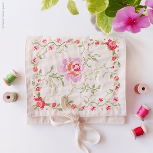 May include: Cream-colored, cross-stitched pouch with a large pink and orange rose in the center, surrounded by a floral border. The pouch is tied with a cream-colored ribbon. Several spools of thread in various colors are scattered around the pouch.