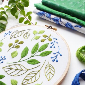 May include: A close-up of a circular embroidery hoop with a white fabric background. The fabric is embroidered with green leaves and blue berries in a floral pattern.
