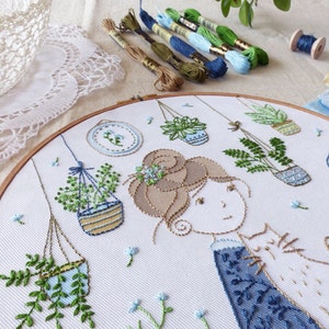 May include: A white embroidery hoop with a design of a woman with brown hair and a cat, surrounded by hanging plants in blue and green pots. The background is white with blue forget-me-not flowers.