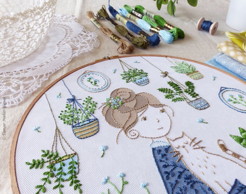 May include: A white embroidery hoop with a design of a woman with brown hair and a cat, surrounded by hanging plants in blue and green pots. The background is white with blue forget-me-not flowers.