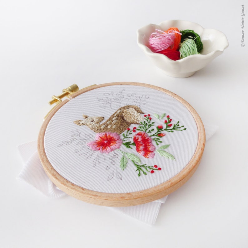 May include: A close-up of a circular embroidery hoop with a white fabric background. The embroidery depicts a brown deer with pink and red flowers and green leaves. The hoop is made of wood and has a gold metal screw.