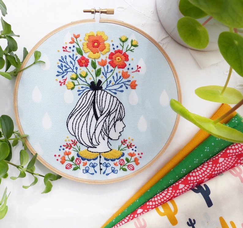 Op de afbeelding: Borduurwerk op een hoepel met een meisje met een bloemenkrans. Het meisje heeft kort, donker haar en draagt een witte kraag. De bloemen zijn kleurrijk en bevatten rood, geel, oranje en roze. De achtergrond is lichtblauw met witte regendruppels.