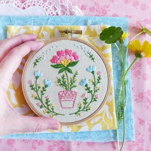 K&ouml;nnte beinhalten: Eine Hand h&auml;lt einen kleinen Stickrahmen mit einer pinkfarbenen Blume, die in der Mitte gestickt ist. Die Blume ist von gr&uuml;nen Bl&auml;ttern und kleineren pinkfarbenen, blauen und gelben Blumen umgeben. Der Rahmen liegt auf einem Stapel Stoff in Gelb, Blau und Pink.