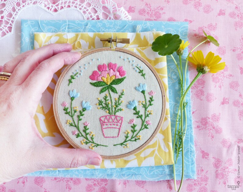 K&ouml;nnte beinhalten: Eine Hand h&auml;lt einen kleinen Stickrahmen mit einer pinkfarbenen Blume, die in der Mitte gestickt ist. Die Blume ist von gr&uuml;nen Bl&auml;ttern und kleineren pinkfarbenen, blauen und gelben Blumen umgeben. Der Rahmen liegt auf einem Stapel Stoff in Gelb, Blau und Pink.