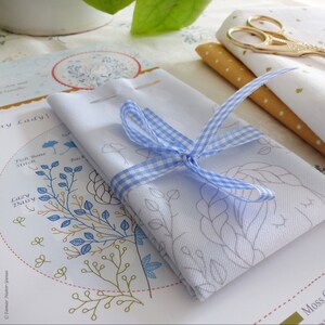 May include: A white fabric with a blue and white gingham ribbon tied around it. The fabric has a floral design printed on it. The fabric is on top of a sheet of paper with instructions for embroidery.
