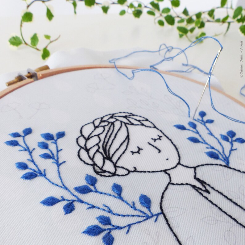 May include: Close-up of a hand embroidery hoop with a blue and black embroidery design of a woman with her eyes closed and blue leaves around her head.