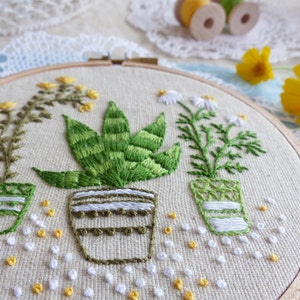 May include: Embroidered hoop art featuring three potted plants: a yellow-flowered plant, a green succulent, and a daisy-like plant. The plants are in green and white striped pots, with white and yellow embroidered accents. The artwork is on a beige fabric.