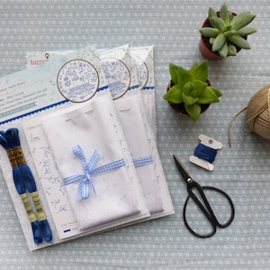 May include: An embroidery kit featuring a white fabric, blue embroidery floss, and a blue and white gingham ribbon. The kit includes instructions for a design of a small house with trees and flowers. The kit is packaged in a white box with the text "tamar" and "Winter Oak" on it.