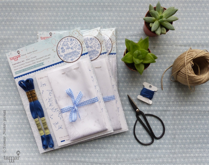 May include: An embroidery kit featuring a white fabric, blue embroidery floss, and a blue and white gingham ribbon. The kit includes instructions for a design of a small house with trees and flowers. The kit is packaged in a white box with the text "tamar" and "Winter Oak" on it.